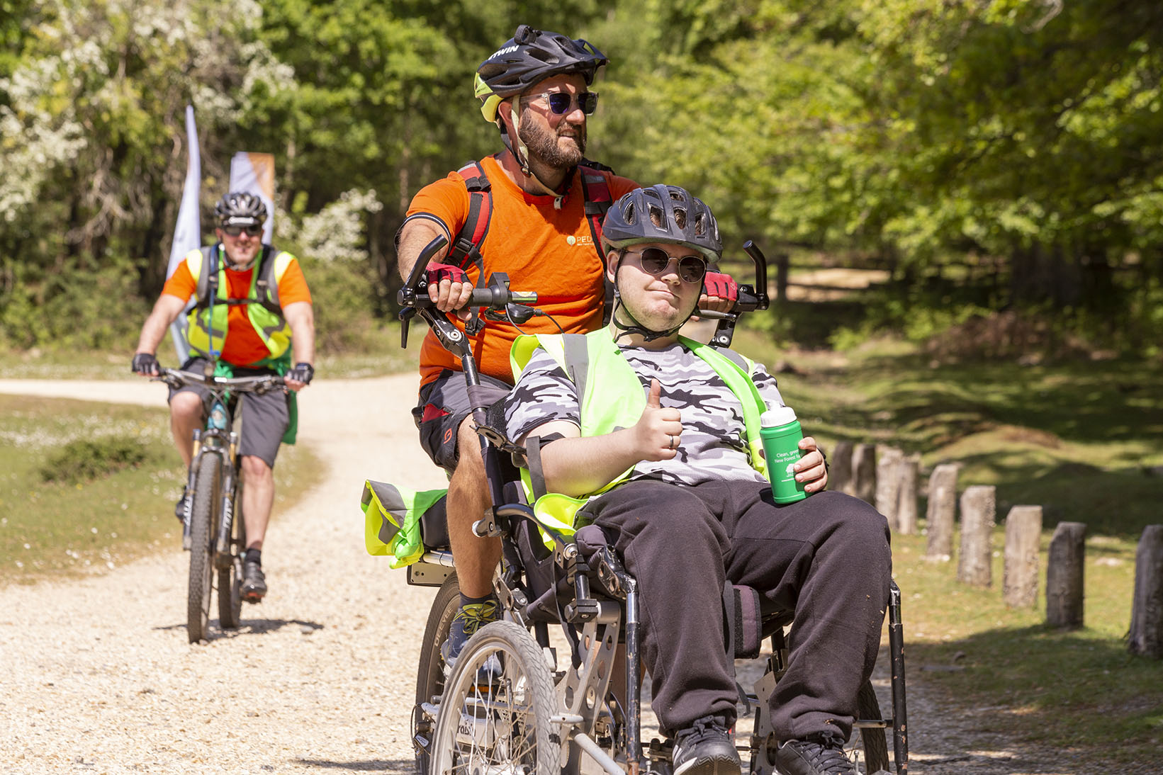 Two cyclists cycling along a summer new forest path with the wheelchair bike passenger giving the camera a thumbs up